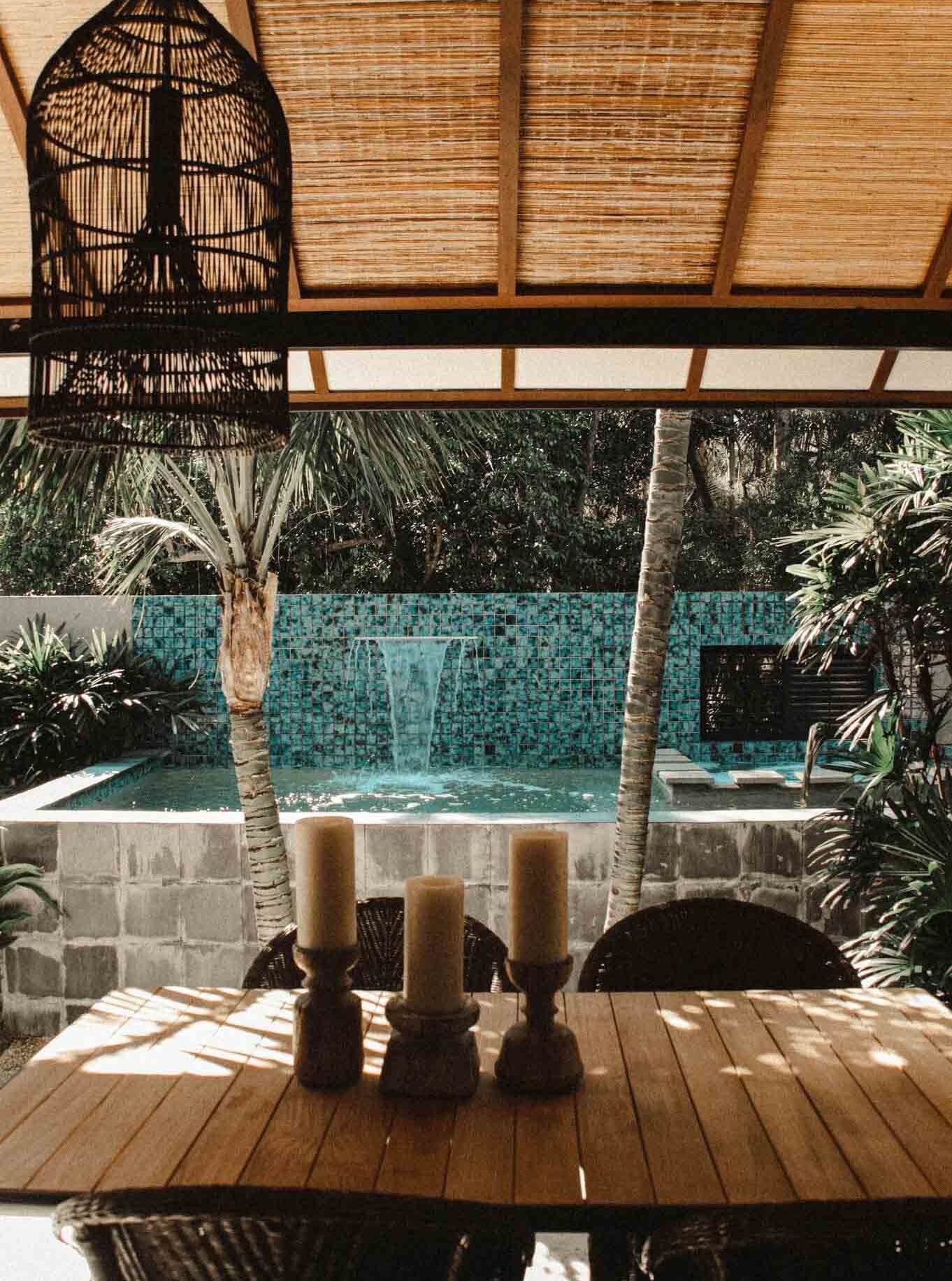 Looking out to the private pool from the undercover verandah with textured pendant light, rustic timber candle holders, comfortable table setting and lush gardens at The Villas of Byron